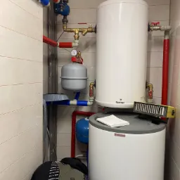 Biały bojler Galmet i Drażice z podłączonymi rurami w kolorach czerwonym i niebieskim, manometr, żółty miernik na górze, w tle płytki ceramiczne.