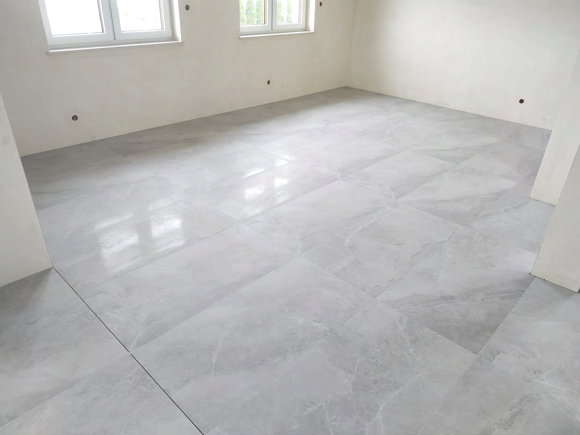 Jasnoszara, marmurkowa posadzka z płytek w nowym pomieszczeniu w Ostrowie Lubelskim, widoczne ślady po szpachlowaniu na ścianach, przygotowanie do dalszych prac wykończeniowych.
