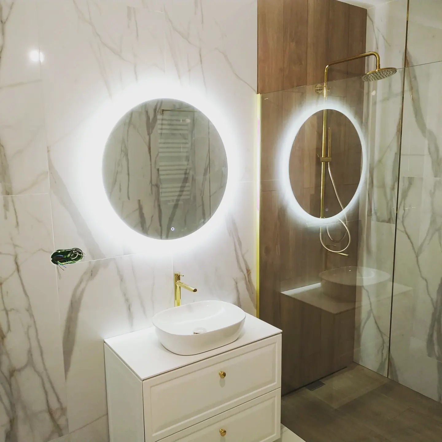 Nowoczesna łazienka z białymi marmurkowymi płytkami, okrągłymi lustrami z podświetleniem LED, białą szafką pod umywalkę z złotymi gałkami i złotą armaturą, widoczne niedokończone podłączenie...