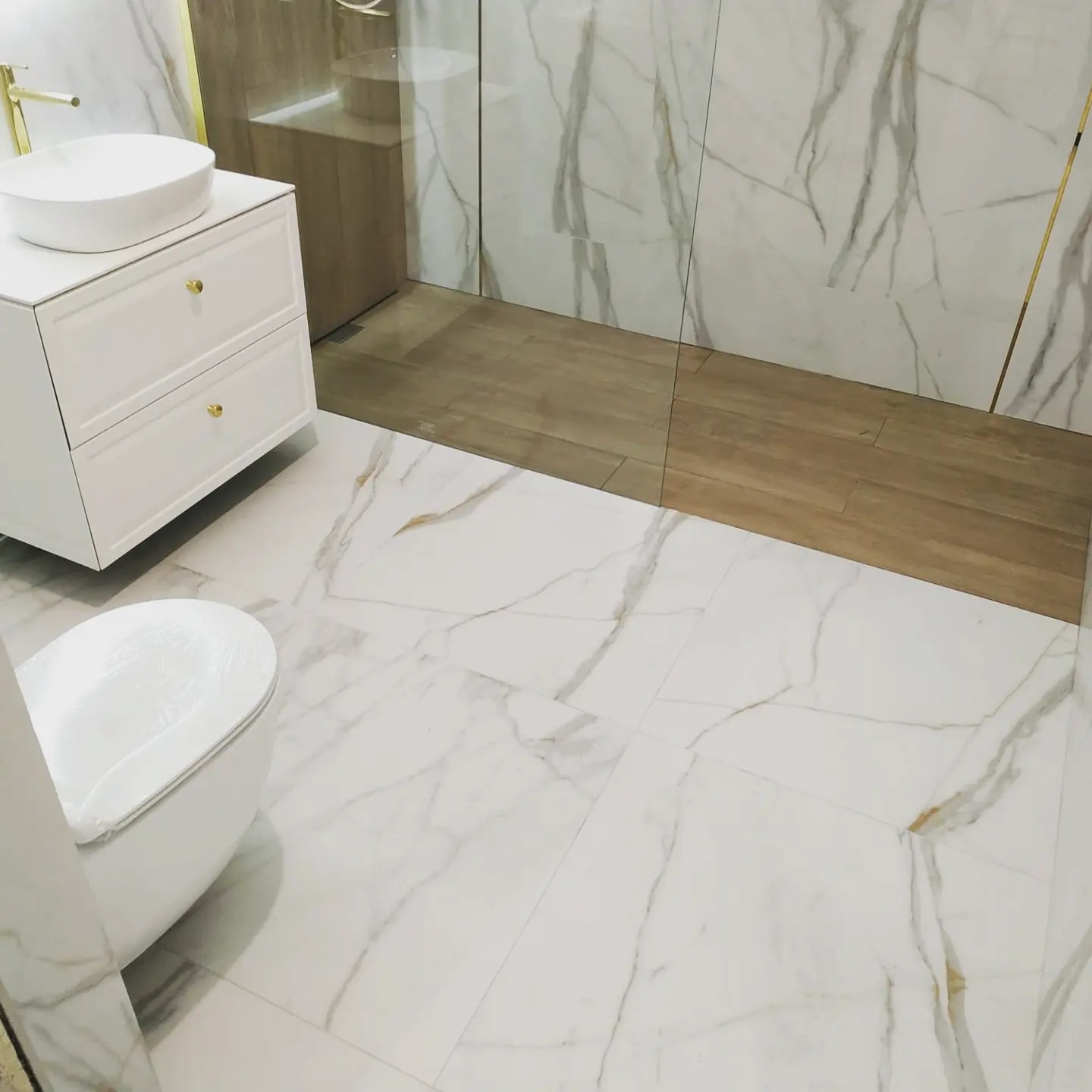 Nowoczesna łazienka z białymi płytkami imitującymi marmur na podłodze i ścianach, drewnopodobną strefą prysznicową, białą szafką pod umywalkę z okrągłą umywalką nablatową oraz toaletą zafoliowaną...