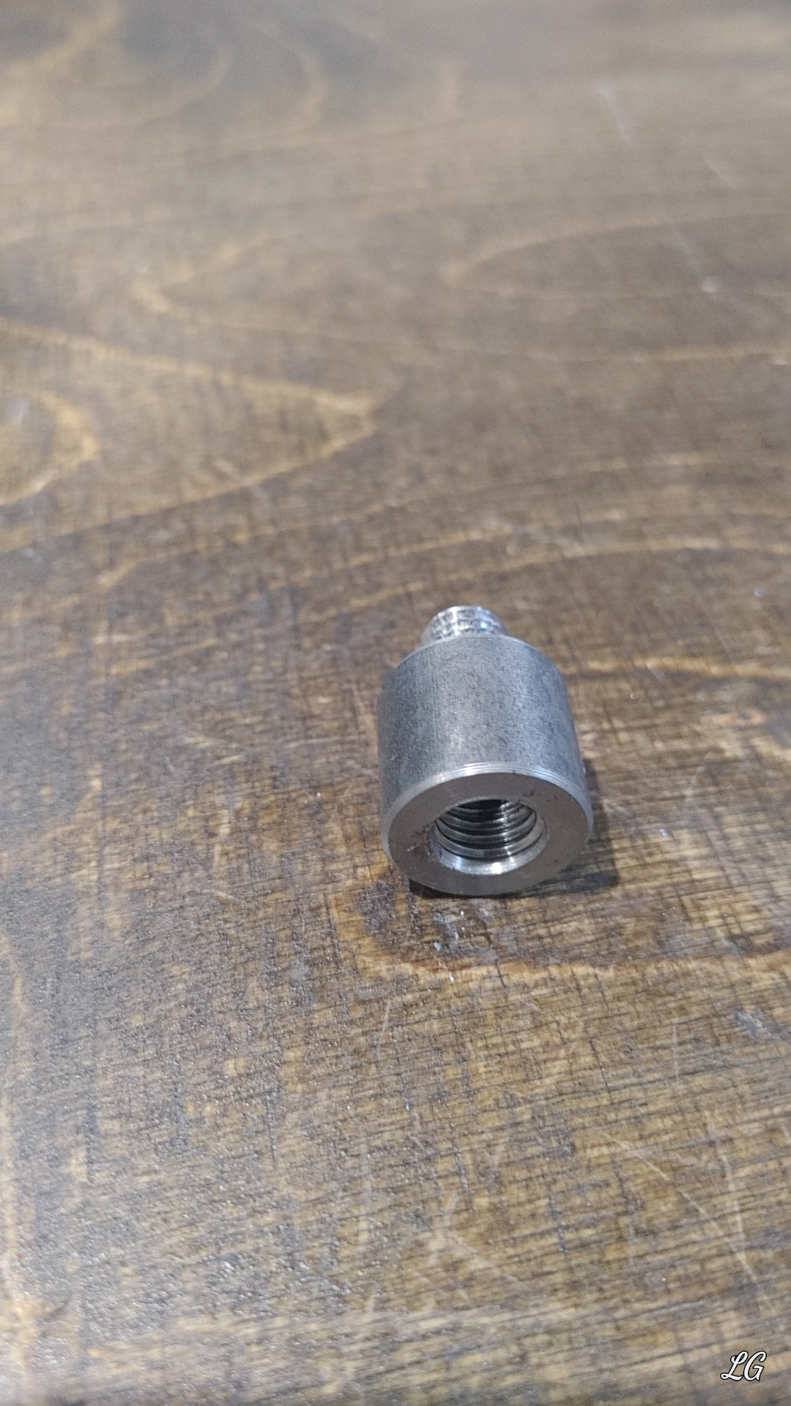 Metalowy element z gwintem wewnętrznym i zewnętrznym, leżący na drewnianym blacie z widocznymi śladami użytkowania.