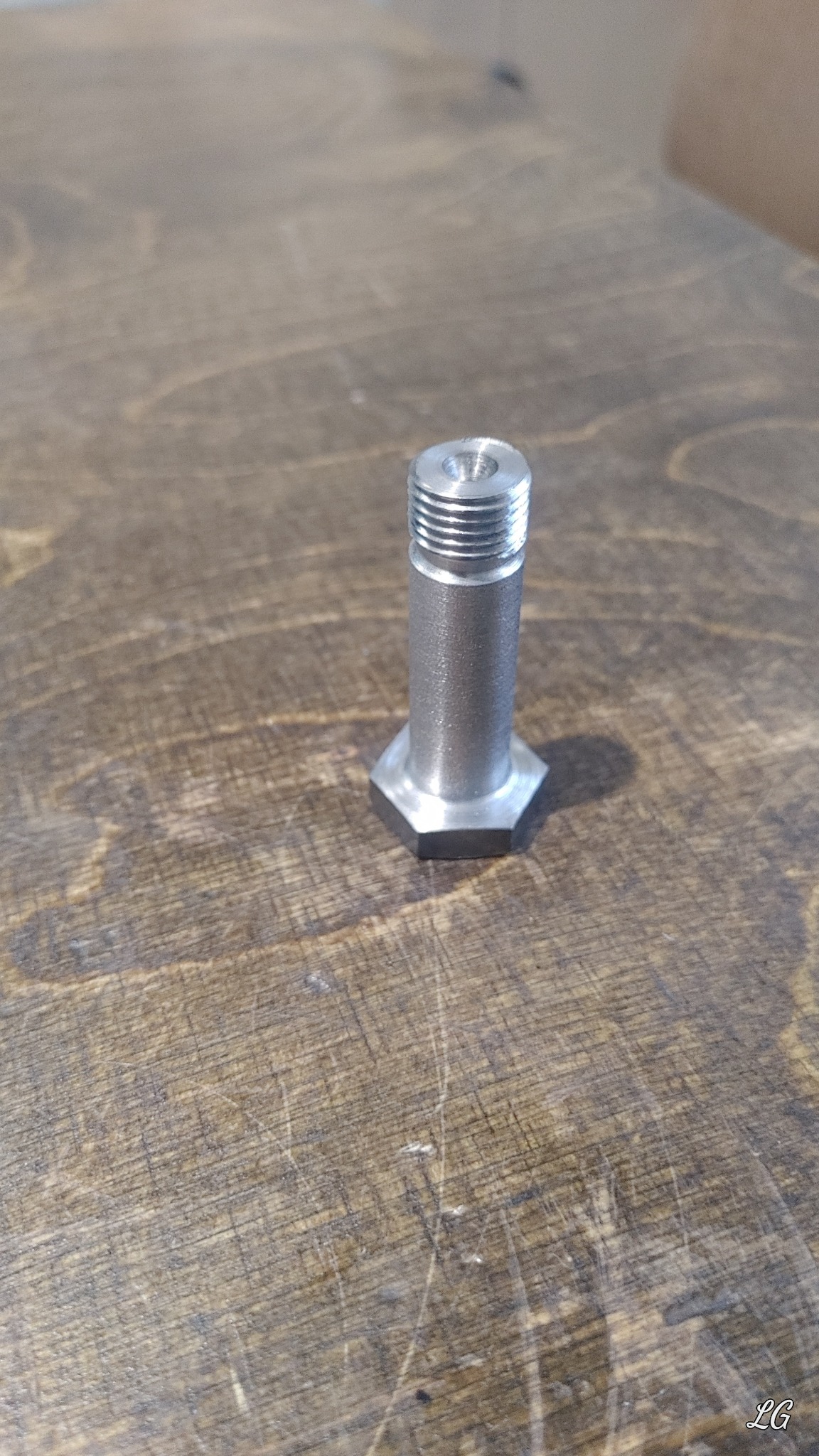 Metalowy trzpień z gwintem i sześciokątną podstawą, obrobiony na tokarce, leżący na drewnianym blacie z widoczną strukturą drewna.