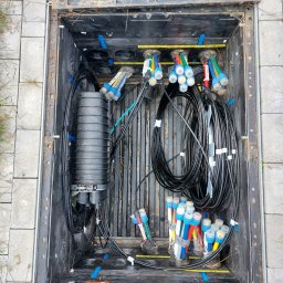 SZM HOLDING SPÓŁKA Z OGRANICZONĄ ODPOWIEDZIALNOŚCIĄ - Otwarta studzienka telekomunikacyjna z kablem światłowodowym i złączkami. Widok z góry na instalację w czarnej skrzynce na tle szarej kostki brukowej.