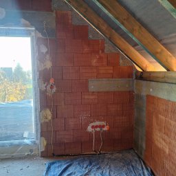 SZM HOLDING SPÓŁKA Z OGRANICZONĄ ODPOWIEDZIALNOŚCIĄ - Wnętrze w trakcie budowy: ściany z czerwonej cegły, okno z widokiem na drzewa, widoczne przewody elektryczne i elementy konstrukcji dachu. Surowy stan.