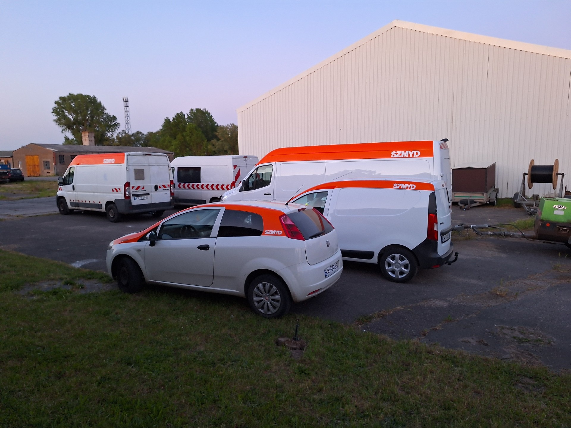 Flota pojazdów firmy SZMYD: biały samochód osobowy i trzy busy z pomarańczowymi akcentami, zaparkowane przed magazynem. Widoczne drzewa i słup telekomunikacyjny w tle.
