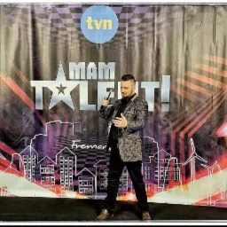 Mężczyzna w ozdobnej marynarce prezentuje się na tle plakatu 'Mam Talent!' z logo TVN, trzymając w ręku rekwizyt.