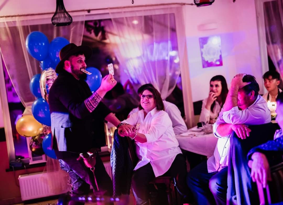 Iluzjonista w kapeluszu prezentuje trik z kartami, trzymając jedną kartę w ustach, podczas występu dla publiczności w udekorowanym sali z balonami.