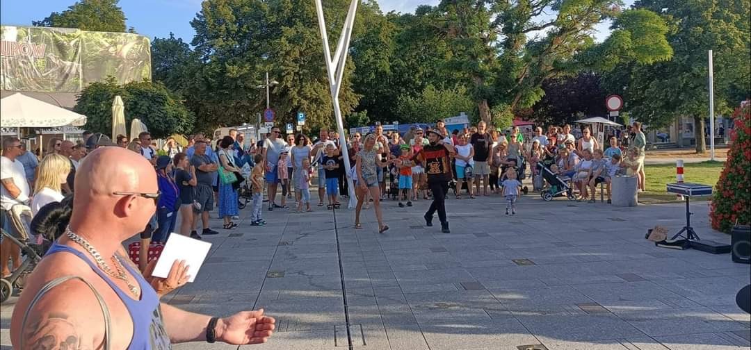 Występ iluzjonisty na placu w Szczecinie, otoczony publicznością, w tle drzewa i budynki, z przodu mężczyzna z kartką.