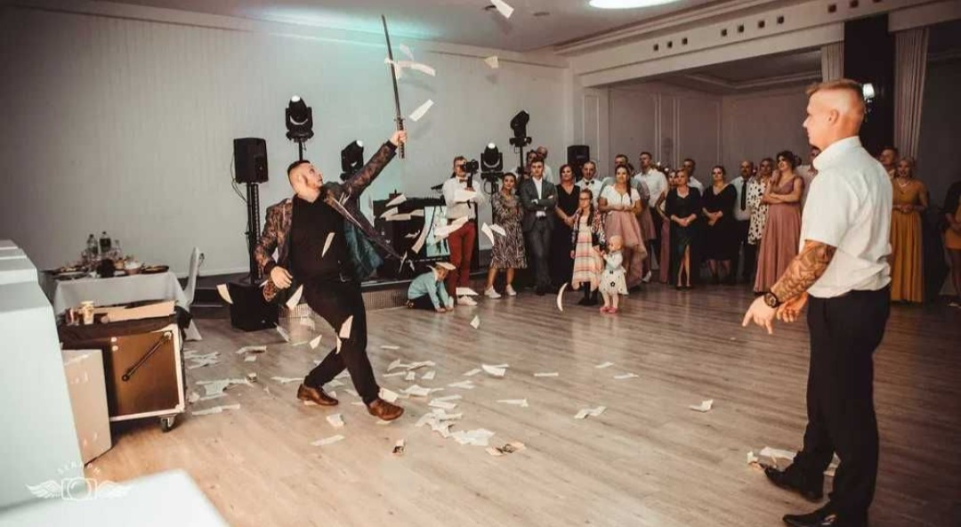 Iluzjonista w trakcie występu na sali, z uniesionym mieczem i rozsypującymi się kartkami, publiczność obserwuje.