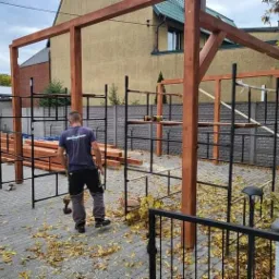 Montaż drewnianej konstrukcji dachu altany ogrodowej, widoczne rusztowania i pracownik z wkrętarką na brukowanym podłożu.