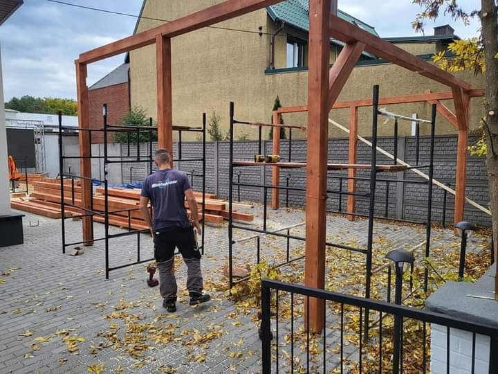 Montaż drewnianej konstrukcji dachu altany ogrodowej, widoczne rusztowania i pracownik z wkrętarką na brukowanym podłożu.