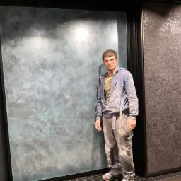 Pracownik w ubraniu roboczym, z plamami farby, prezentuje ścianę z dekoracyjną, turkusowo-szarą gładzią w stylu marmuru, obok ściana z ciemną gładzią z brokatem.