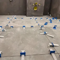 Szare płytki podłogowe w trakcie układania, z widocznymi systemami poziomowania płytek (klipsy i kliny) z niebieskimi elementami, na tle surowych ścian w szarym kolorze i widocznymi fragmentami...