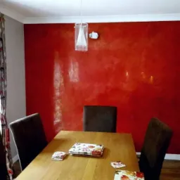 Ściana w jadalni pomalowana na intensywny czerwony kolor z efektem dekoracyjnym, widoczne krzesła i stół z nakryciem, lampa wisząca nad stołem, okno z zasłoną po lewej stronie.