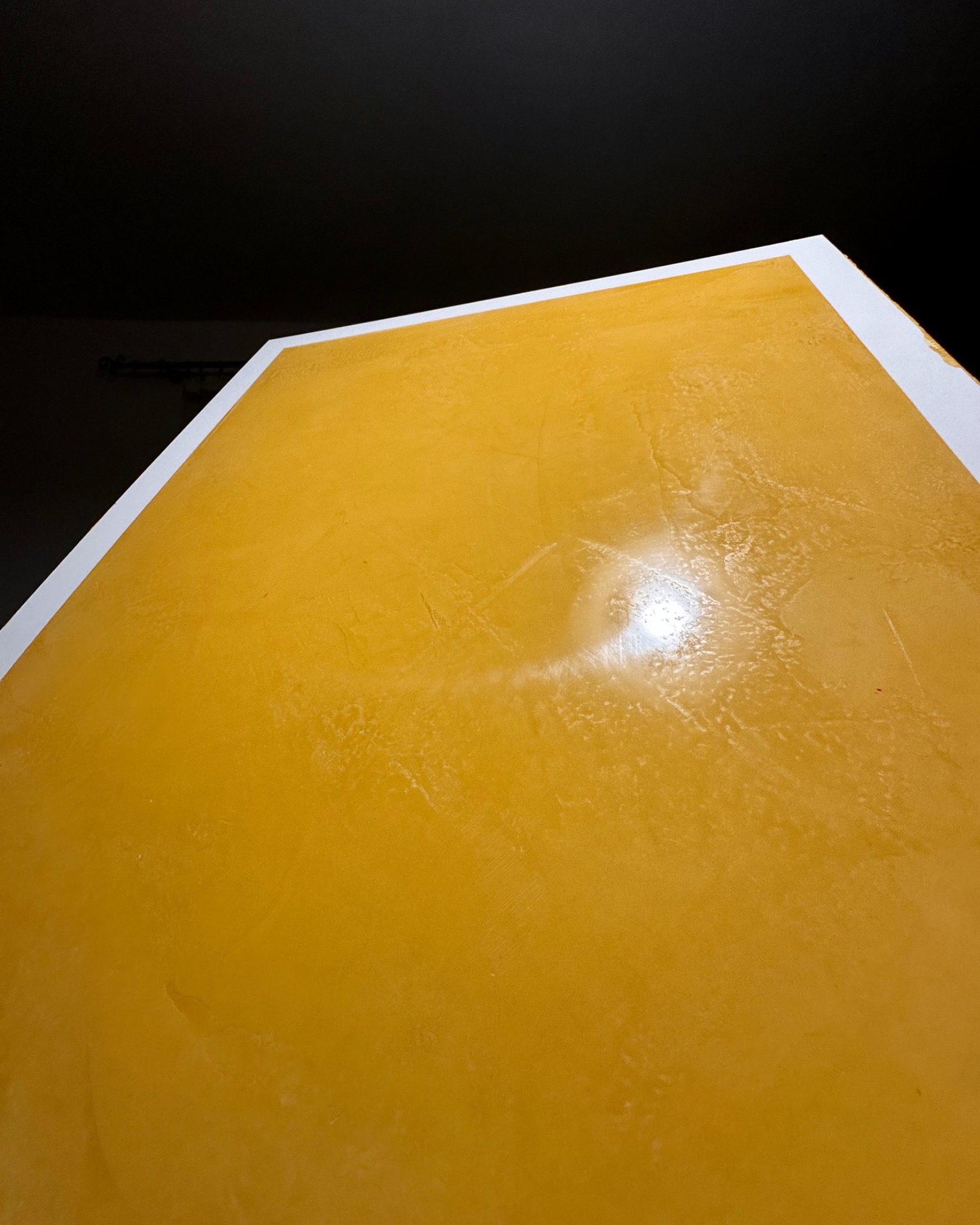 Żółta, nierówna powierzchnia ściany z białym obramowaniem pod kątem, oświetlona punktowo, z ciemnym tłem.