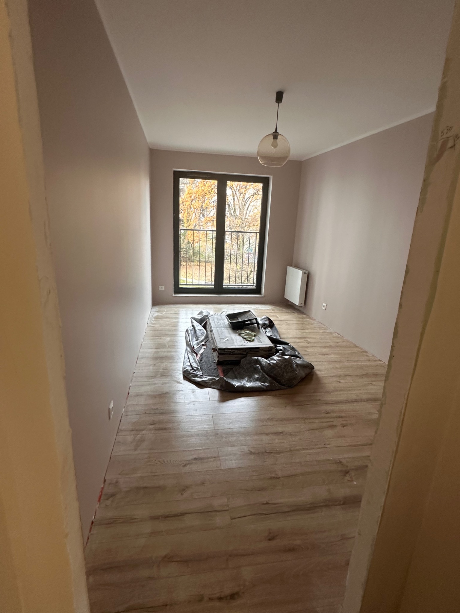 Pokój po położeniu paneli podłogowych w odcieniach drewna, ściany pomalowane na jasny róż, okno balkonowe z widokiem na drzewa jesienią, zabezpieczone folią malarską narzędzia na środku pomieszczenia.