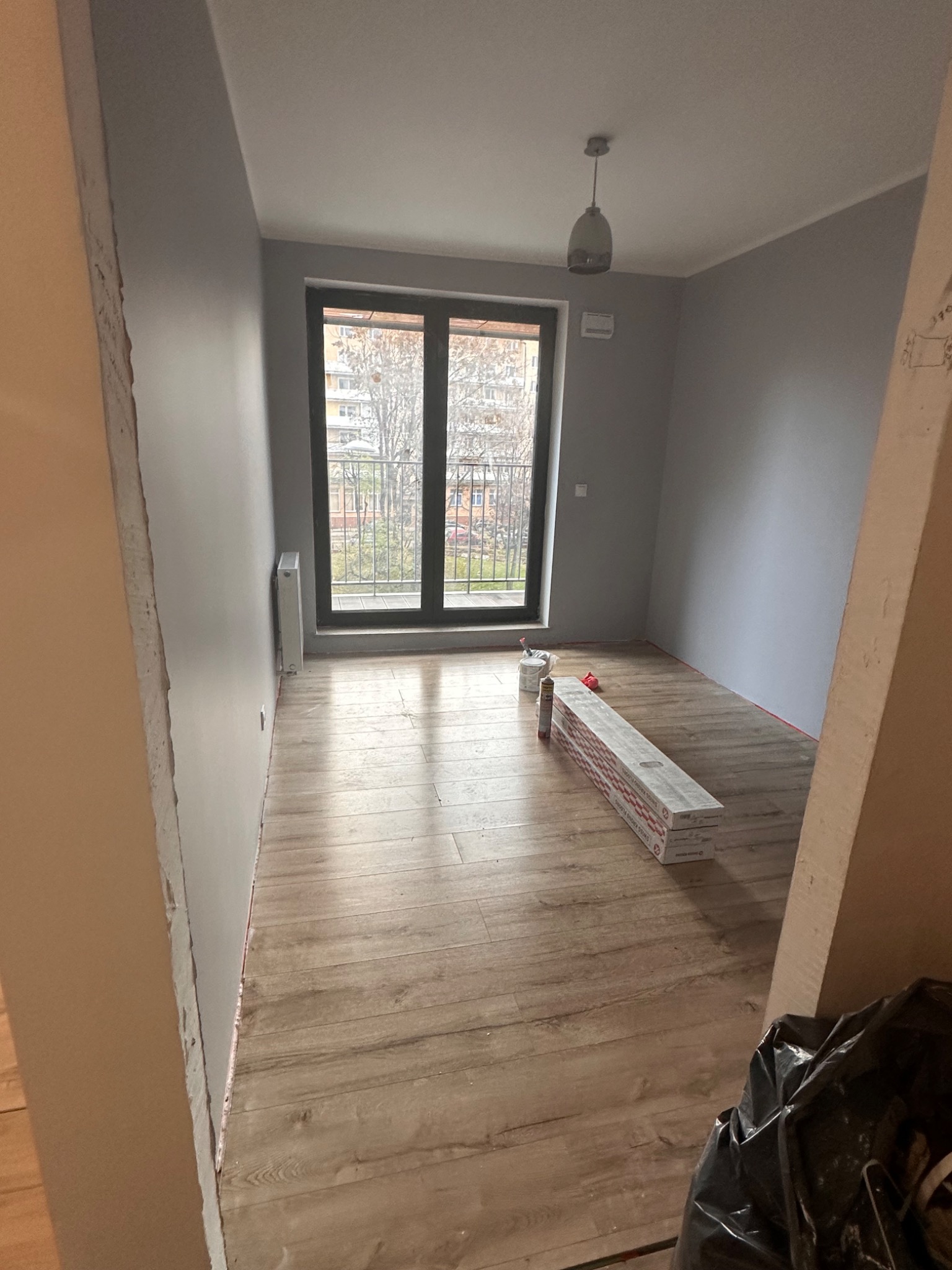 Pokój w trakcie prac wykończeniowych: świeżo pomalowane szare ściany, ułożona podłoga z paneli, paczki paneli leżące na podłodze, widok na balkon przez okno.