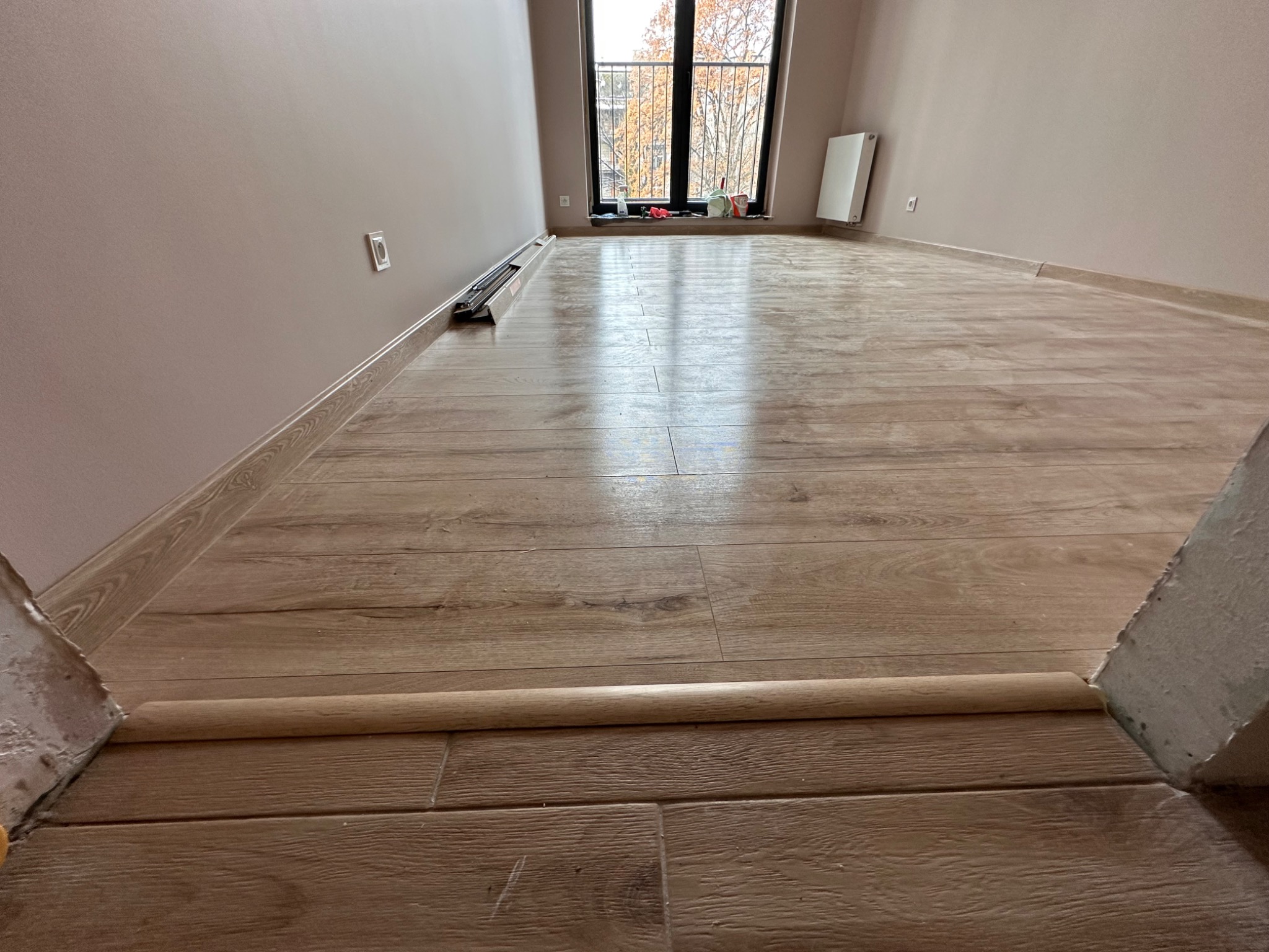 Nowo ułożone panele podłogowe z drewnopodobnym wzorem, widoczne listwy przypodłogowe i próg, surowe ściany w jasnym odcieniu beżu, okno z widokiem na drzewa bez liści.