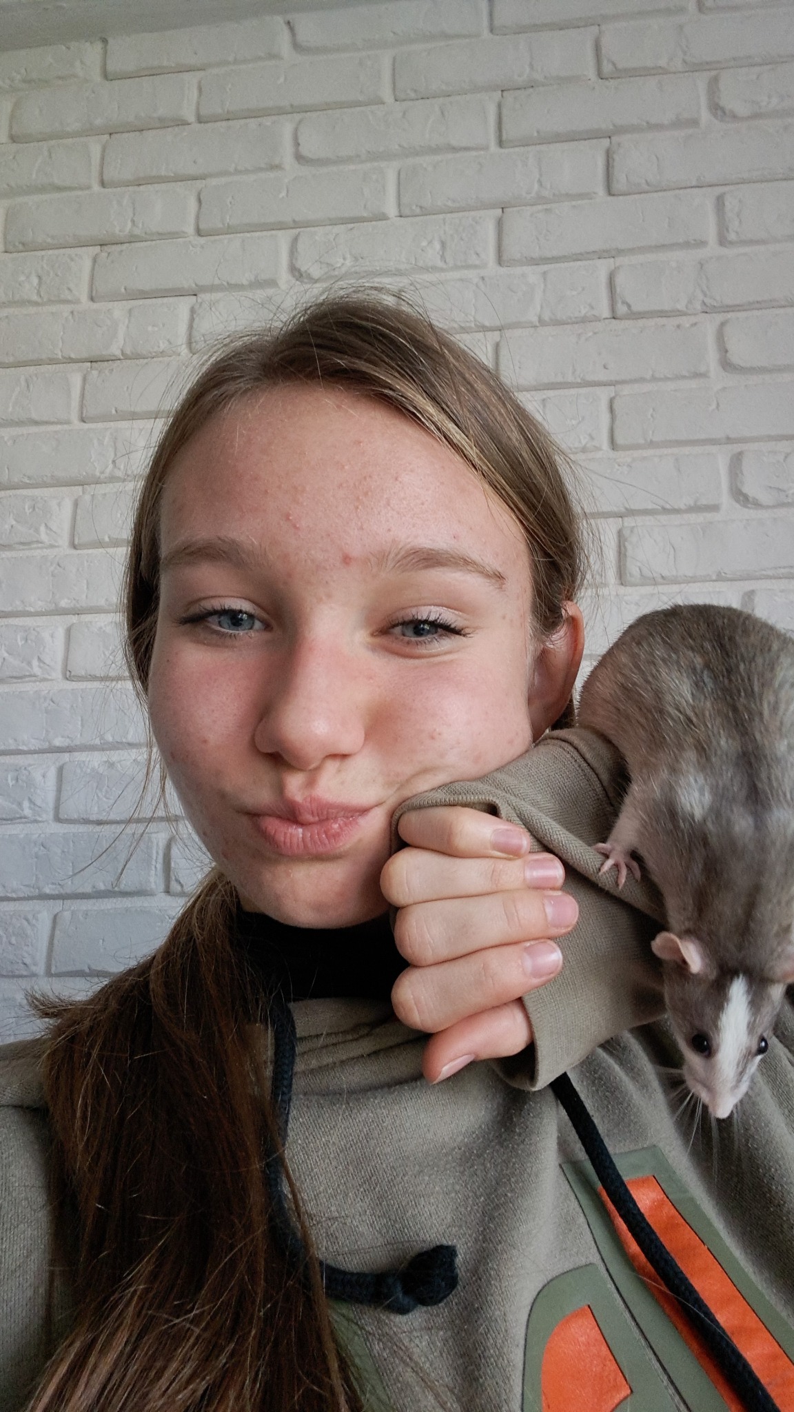 Młoda kobieta z niebieskimi oczami i zaczerwienioną cerą trzyma na ramieniu szarego szczura z białym pyskiem na tle białej ściany z cegieł.