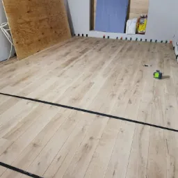 Układanie jasnych paneli podłogowych z widocznymi czarnymi liniami wyznaczającymi obszar roboczy, poziomica laserowa na podłodze, płyta kartonowo-gipsowa oparta o ścianę, worki z zaprawą w tle.