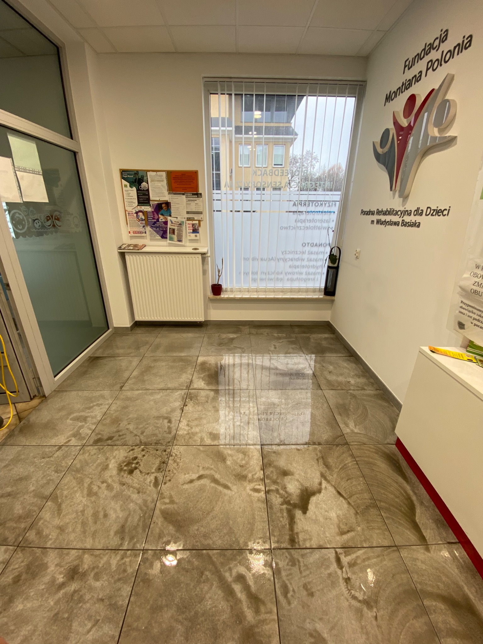 Mokre, umyte płytki podłogowe w jasnym pomieszczeniu z oknem, widoczny radiator i tablica ogłoszeń na ścianie, odbicie światła na podłodze.