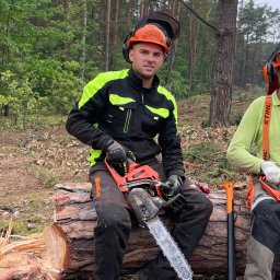 Michał Szyszka - Młody mężczyzna w kasku i kurtce roboczej siedzi na ściętym pniu w lesie, trzymając piłę spalinową Stihl; w tle widać drzewa i drugiego pracownika z siekierą.