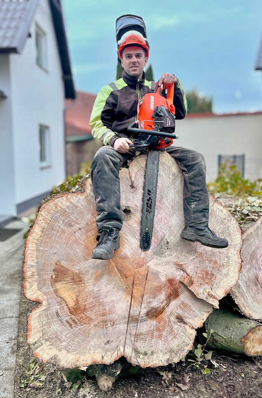 Drwal w hełmie siedzi na pniu drzewa z piłą spalinową Echo, widoczne ślady cięcia na pniu.