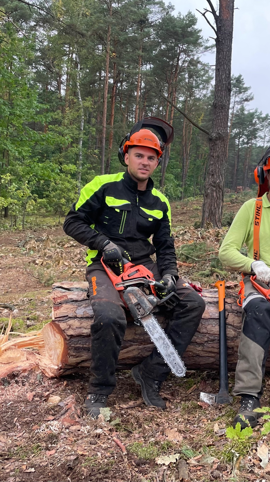 Młody mężczyzna w kasku i kurtce roboczej siedzi na ściętym pniu w lesie, trzymając piłę spalinową Stihl; w tle widać drzewa i drugiego pracownika z siekierą.