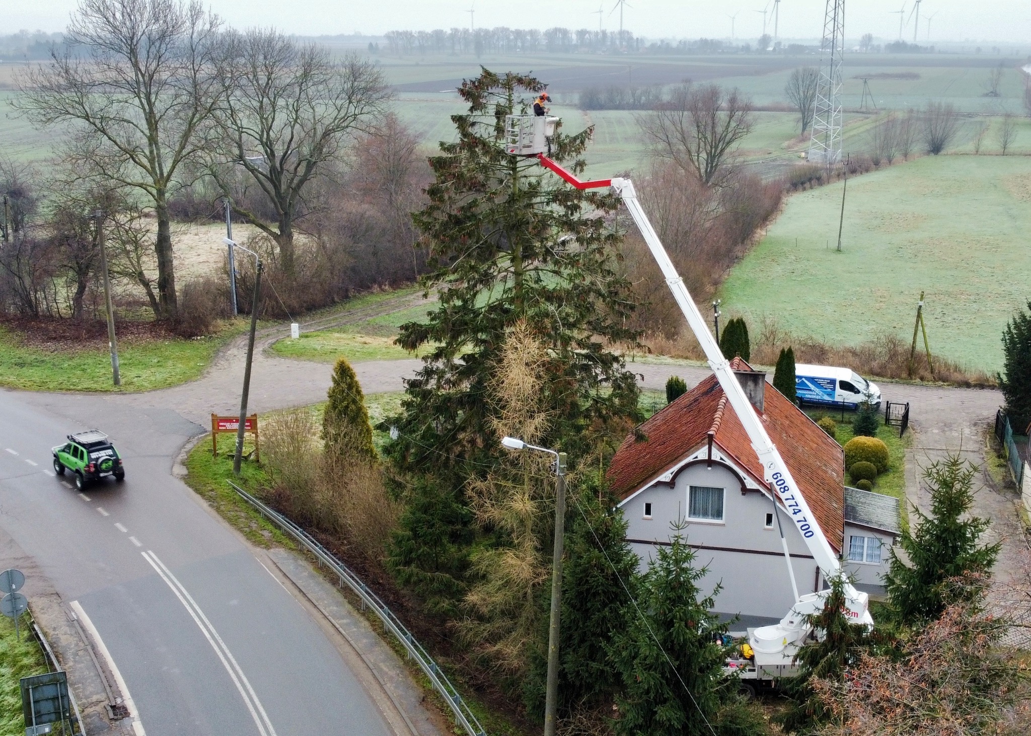 Pracownik w podnośniku koszowym przycina wysokie drzewo iglaste w Nowym Dworze Gdańskim, widoczny dom z czerwoną dachówką, oraz zielony samochód terenowy na ulicy.