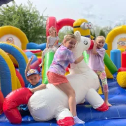 Dzieci bawią się na dmuchanym placu zabaw z motywem Minionków, w tym biały dmuchany jednorożec z czerwonymi detalami, na którym siedzi dwoje dzieci.