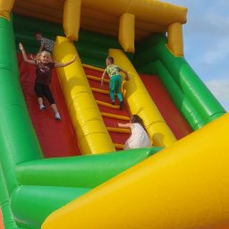 P.W. Mieczysław Grzeszczuk - Dzieci bawiące się na dmuchanej zjeżdżalni w kolorach żółtym, zielonym, czerwonym i niebieskim, z widocznym niebem w tle.