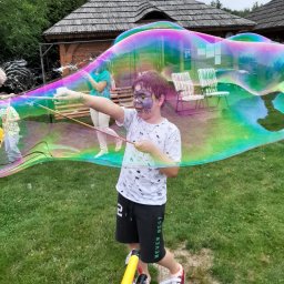 P.W. Mieczysław Grzeszczuk - Chłopiec z pomalowaną twarzą tworzy ogromną tęczową bańkę mydlaną za pomocą specjalnych kijów na trawniku, w tle drewniane budynki.