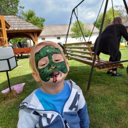 P.W. Mieczysław Grzeszczuk - Chłopiec z pomalowaną twarzą na zielono, imitującą maskę potwora z wystawionym czerwonym językiem, na tle trawnika z huśtawką i altaną.