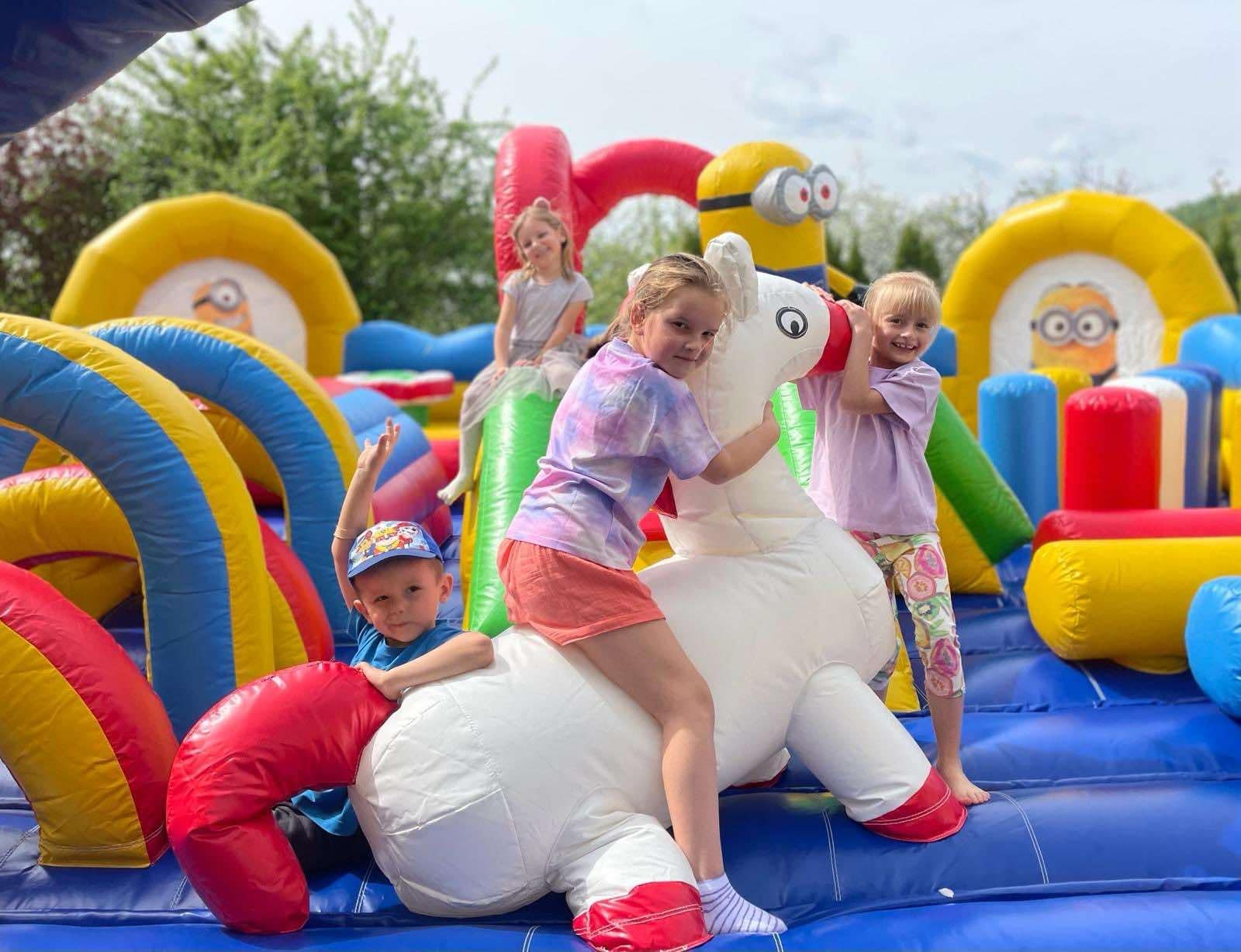 Dzieci bawią się na dmuchanym placu zabaw z motywem Minionków, w tym biały dmuchany jednorożec z czerwonymi detalami, na którym siedzi dwoje dzieci.