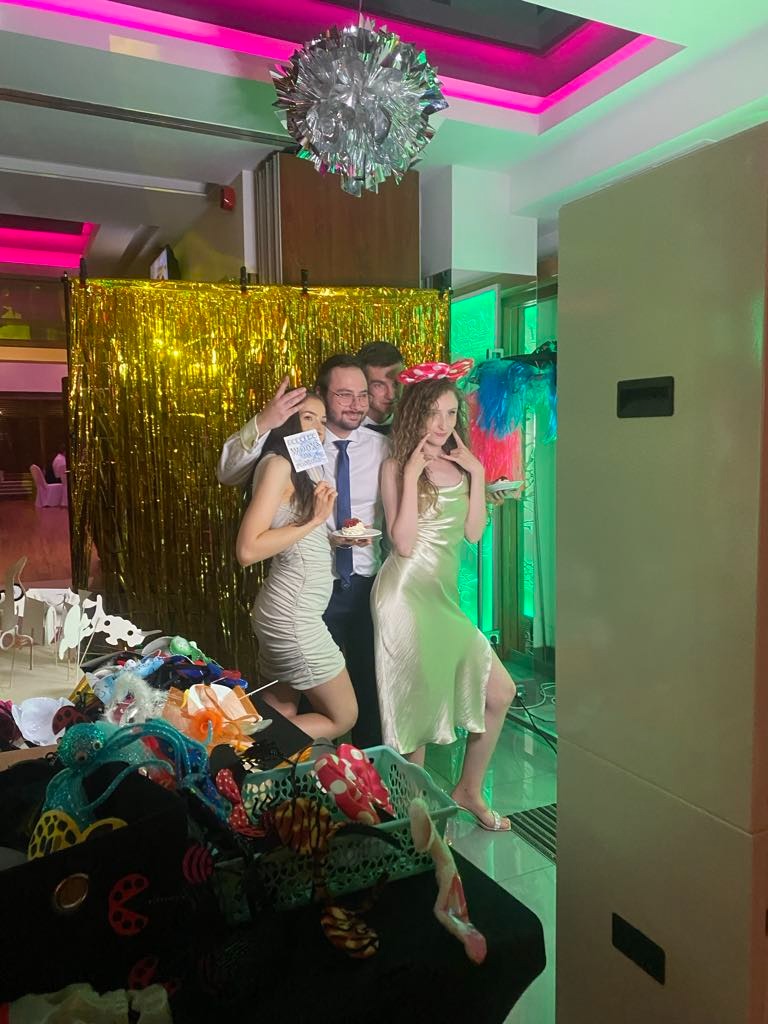 Grupa czterech osób pozuje do zdjęcia przed złotą, błyszczącą ścianką. Dwie kobiety w sukienkach i dwóch mężczyzn, jeden w koszuli i krawacie, drugi w czapce. W koszyku leżą rekwizyty do zdjęć.