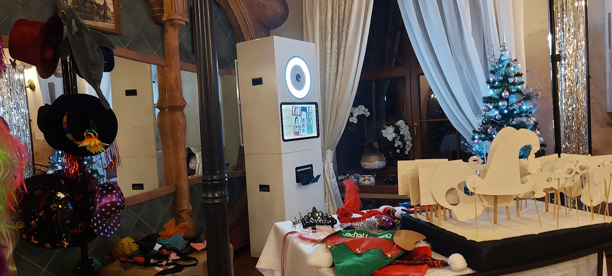 Biała kabina z ekranem i oświetleniem kołowym, otoczona rekwizytami takimi jak kapelusze, peruki i maski, ustawiona w pomieszczeniu z oknem i choinką w tle.