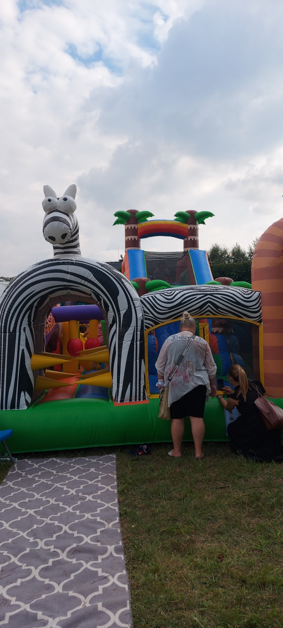 Duży, dmuchany zamek w kształcie zebry i dżungli, z dwoma osobami dorosłymi w pobliżu, ustawiony na trawie obok wzorzystego dywanu.