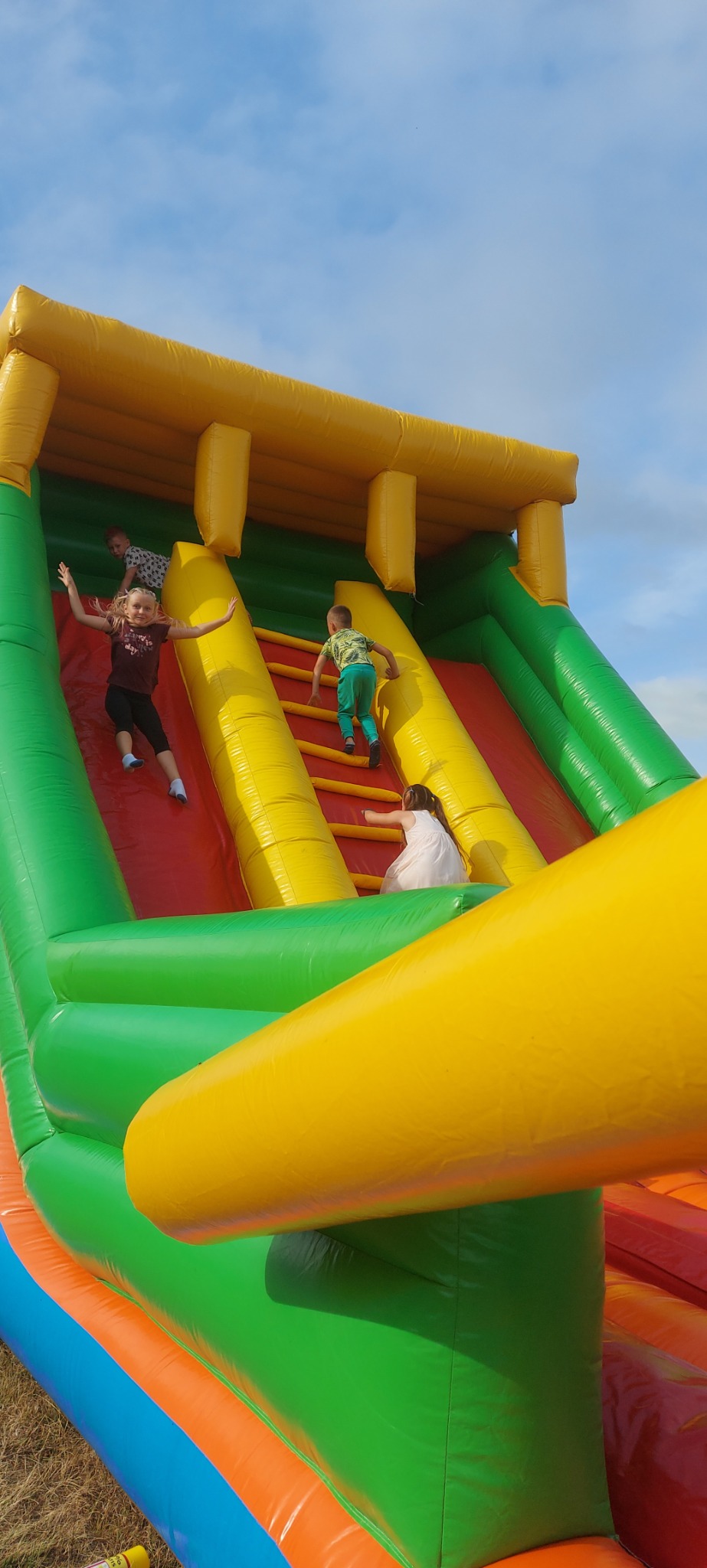 Dzieci bawiące się na dmuchanej zjeżdżalni w kolorach żółtym, zielonym, czerwonym i niebieskim, z widocznym niebem w tle.