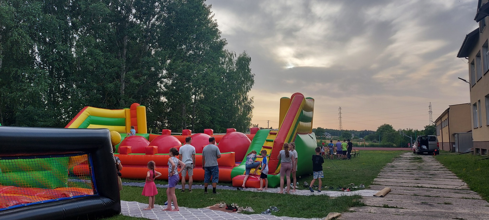 Rozłożone dmuchańce na trawie, z bawiącymi się dziećmi i dorosłymi, w tle budynek i drzewa, pochmurne niebo.
