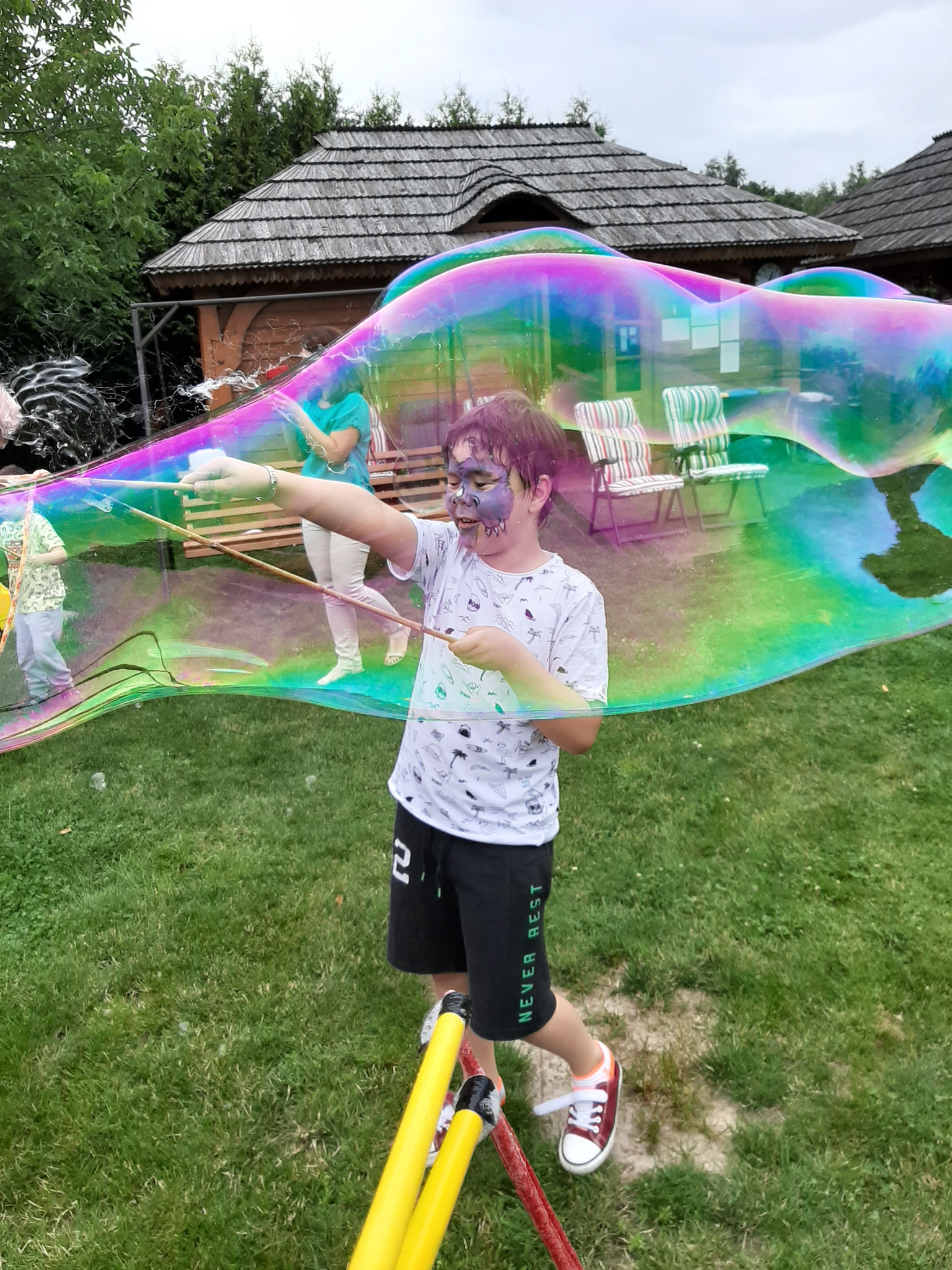 Chłopiec z pomalowaną twarzą tworzy ogromną tęczową bańkę mydlaną za pomocą specjalnych kijów na trawniku, w tle drewniane budynki.
