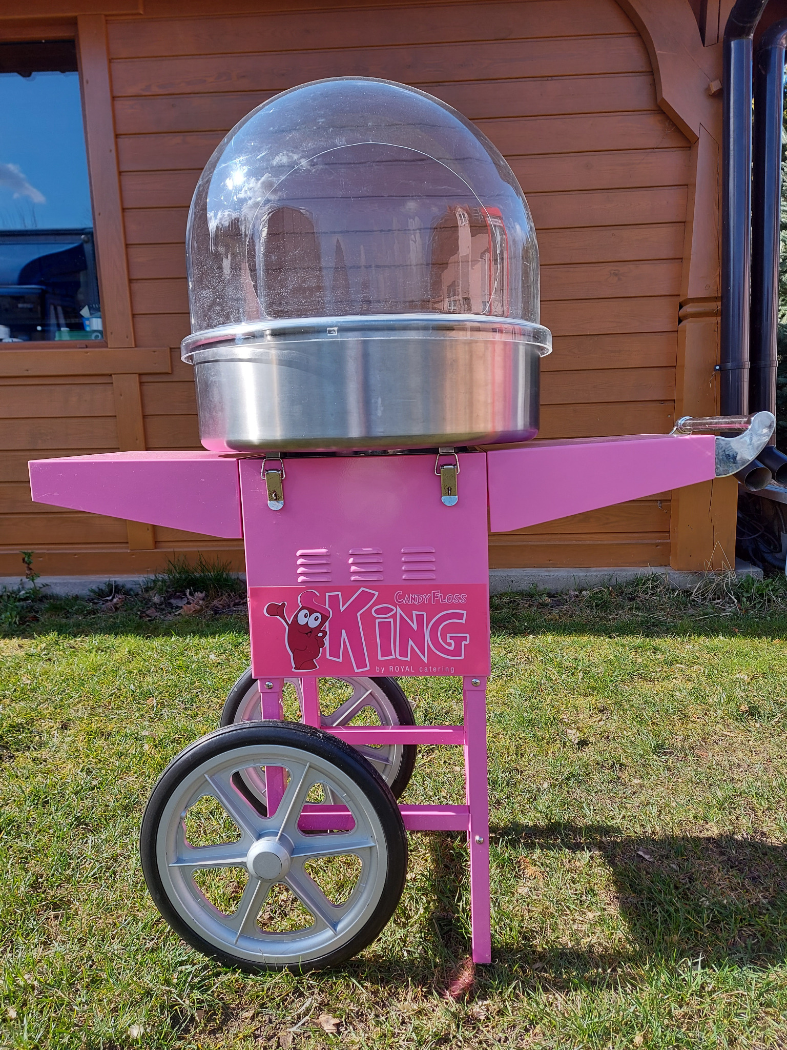 Różowy wózek do waty cukrowej z przezroczystą kopułą i logo 'King Candy Floss by ROYAL catering' na tle drewnianego budynku i trawnika.