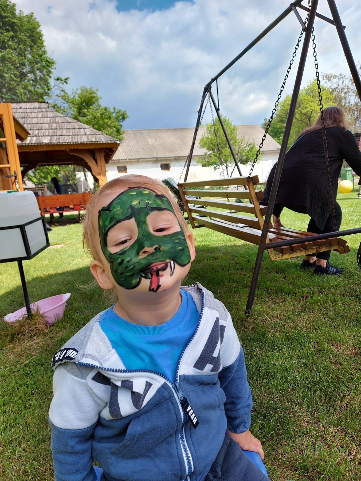 Chłopiec z pomalowaną twarzą na zielono, imitującą maskę potwora z wystawionym czerwonym językiem, na tle trawnika z huśtawką i altaną.