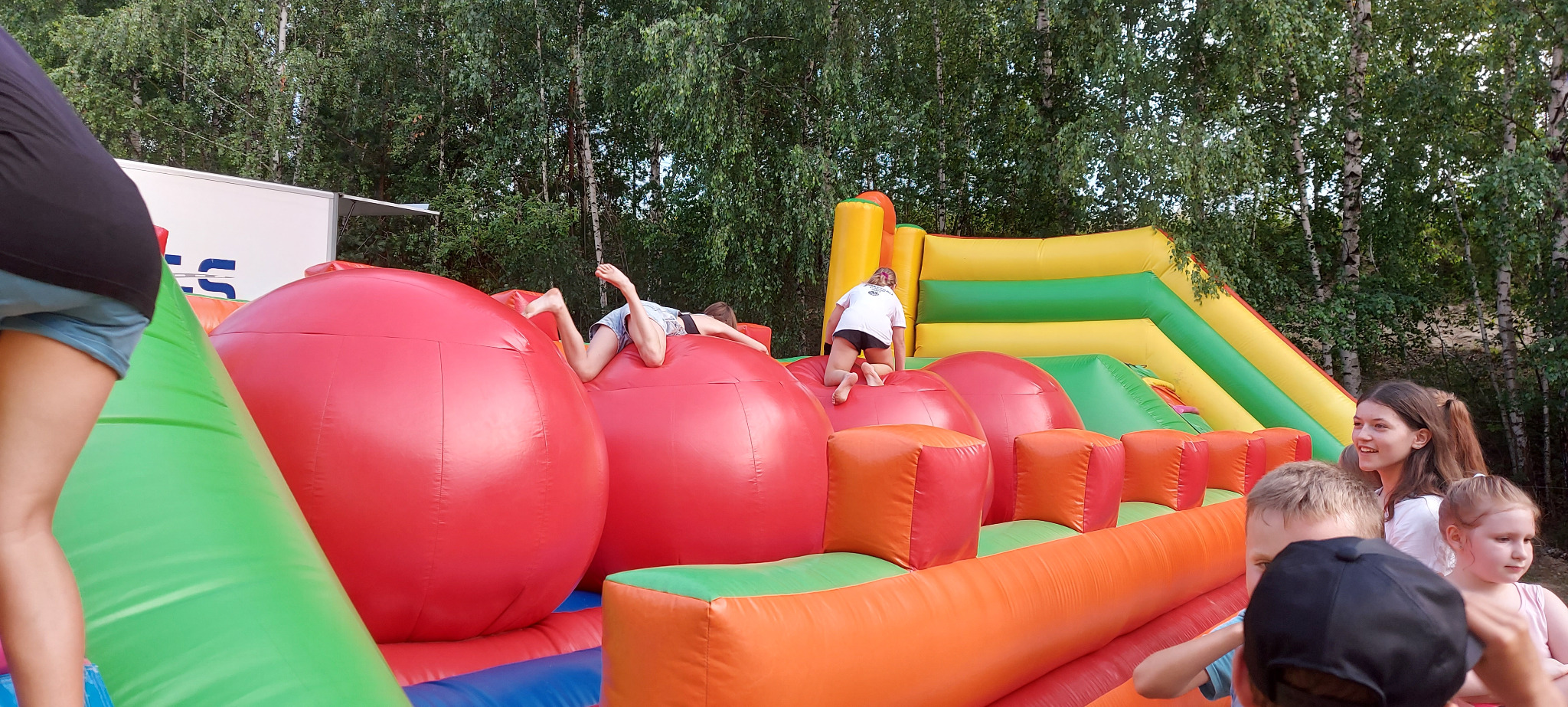 Dzieci bawią się na dmuchanym torze przeszkód z kulami i zjeżdżalnią, z widocznymi drzewami w tle.