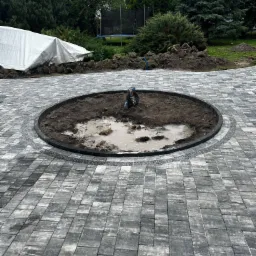 Szara kostka brukowa ułożona wokół okrągłego wykopu z błotnistą wodą, przygotowanego pod nasadzenia, z widocznym zwiniętym wężem ogrodowym. W tle trawnik i zieleń.