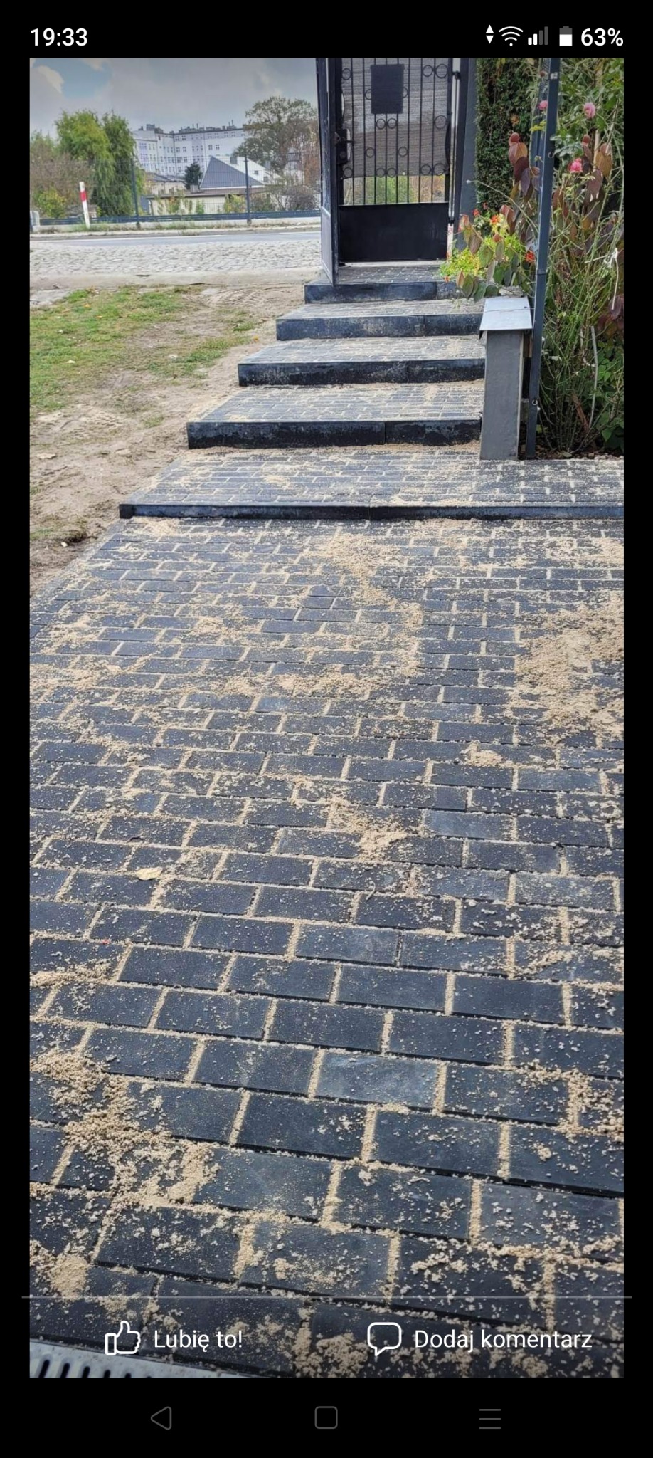 Nowo ułożona, ciemnoszara kostka brukowa na podjeździe, posypana piaskiem kwarcowym, prowadząca do schodów wejściowych z metalową bramką i zielenią po bokach.