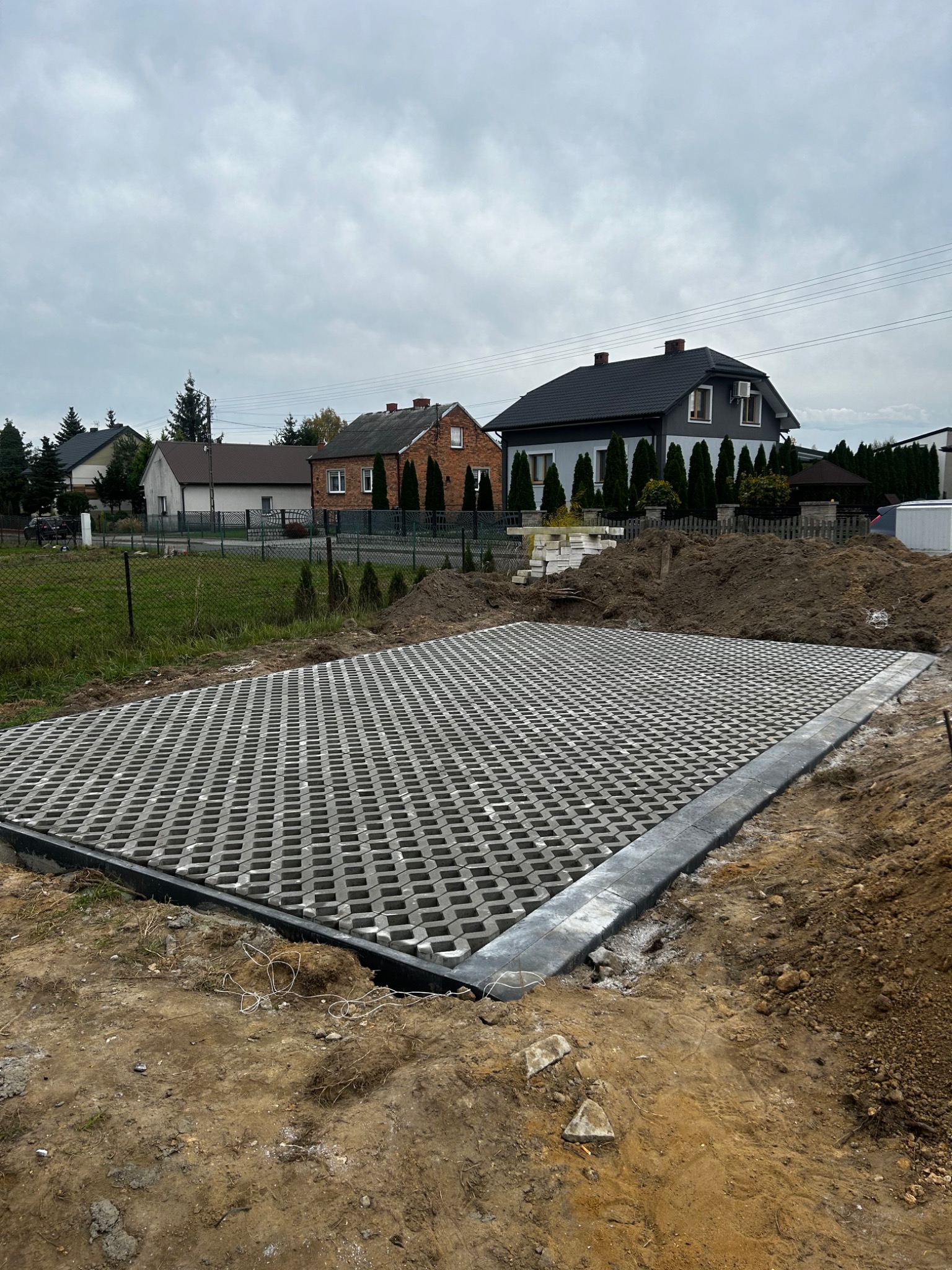 Ułożona, szara ażurowa kostka brukowa na przygotowanym podłożu, widoczne obrzeża, w tle domy jednorodzinne i pochmurne niebo.