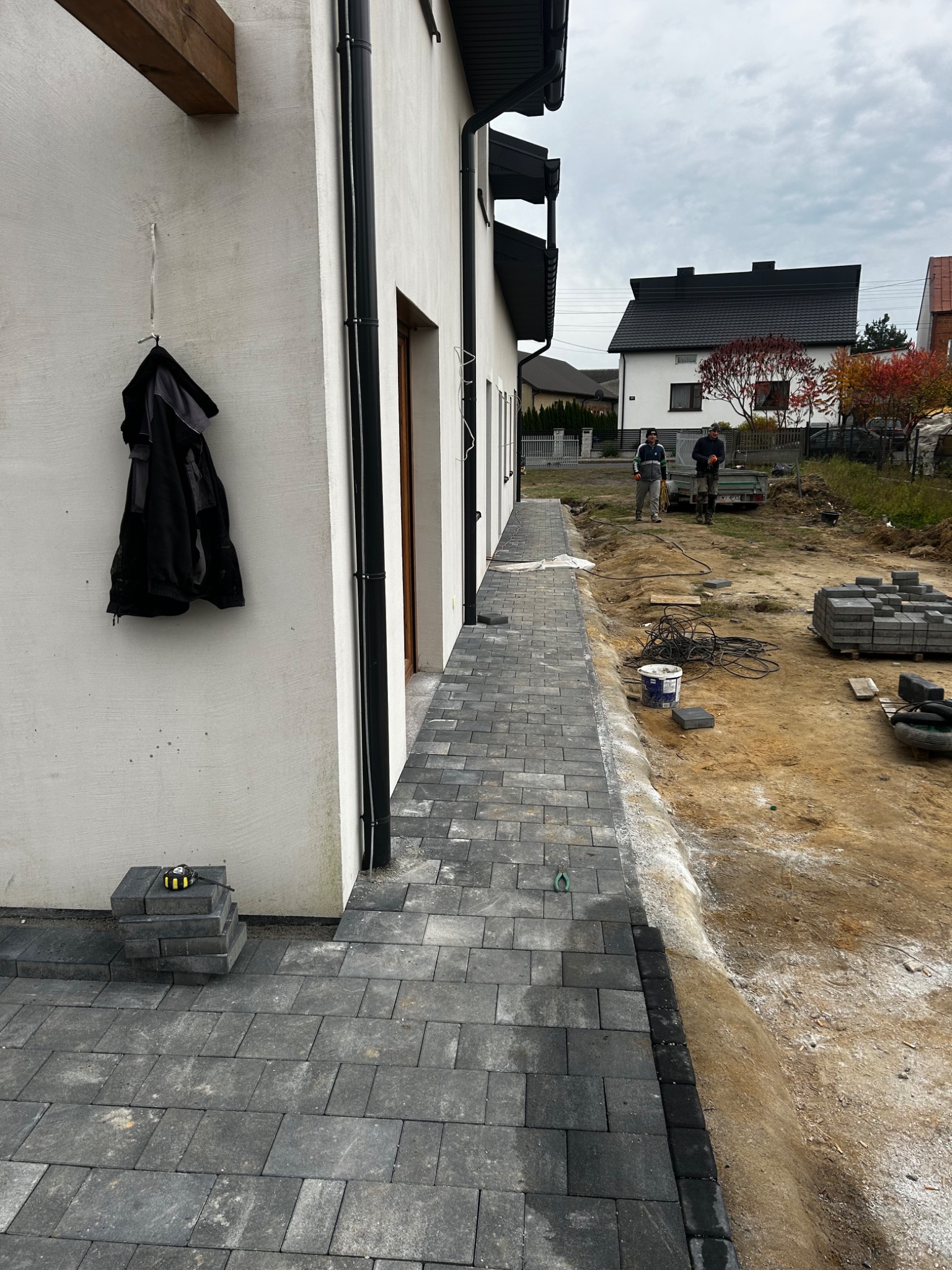 Ułożona ścieżka z szarej kostki brukowej przy białym budynku w Widoradzu Górnym. W tle widoczni pracownicy i przygotowany teren pod dalsze prace.