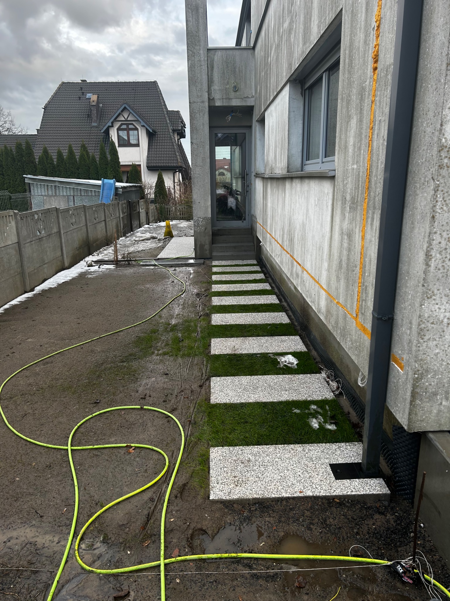 Ścieżka z kwadratowych płyt ułożonych na trawie prowadząca do wejścia budynku, widoczny fragment elewacji z oknem, na ziemi leży zwinięty, zielony wąż ogrodowy, w tle dom jednorodzinny.