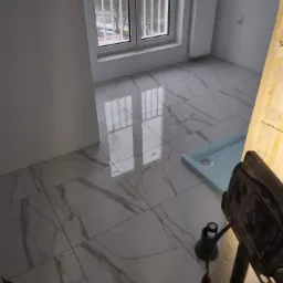 Błyszczące, marmurowe płytki podłogowe w trakcie układania w jasnym pomieszczeniu z oknem i grzejnikiem. Widoczny brodzik oraz narzędzia glazurnicze.