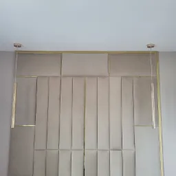 Beżowy panel ścienny z aksamitną fakturą, zdobiony złotymi listwami, oświetlony wiszącymi lampami z elementami złota.